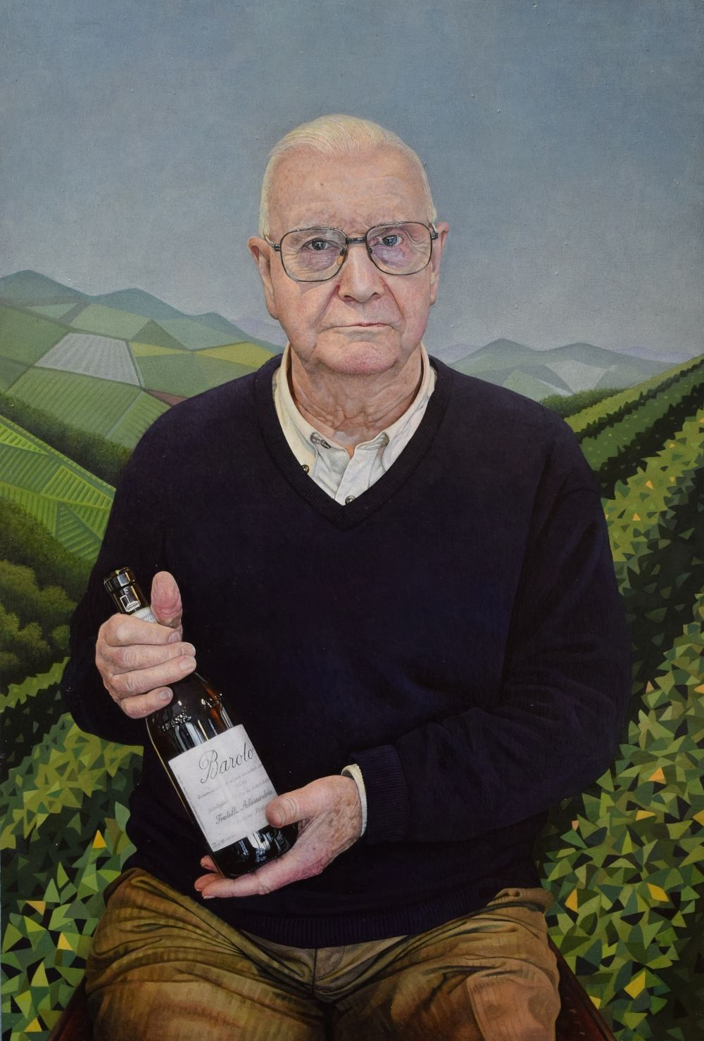 A man seated looking at the viewer holding a bottle of Barola wine with vineyards in the background