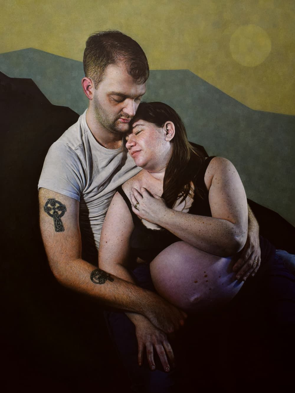 A man and a woman embracing. The man has a tattoo on his arm and the woman is heavily pregnant.